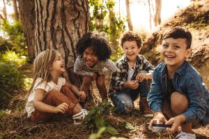 Kinder sitzen lachend zusammen im Wald vor einem Baum.