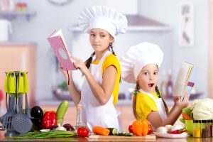Zwei Kinder mit Kochmütze und Schürze schauen mit dem Rücken zu einander in Kochbücher in der Küche..