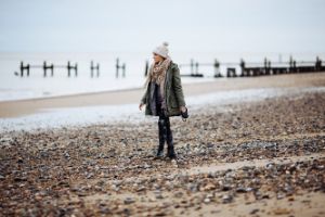 Eine Frau mit Jacke, Schal und Mütze geht am Strand spazieren.