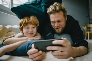 Ein Vater liegt mit seinem Sohn auf dem Bett mit vielen Kissen und schauen lächelnd auf ein Smartphone.